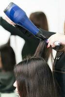 Hands of beautician drying long brunette hair of client using blue hairdryer and hairbrush in salon photo