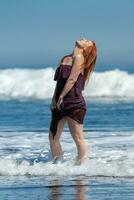 Happy woman in long dress with raised hem standing ankle deep in water on beach, head looking upward photo
