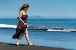 pelirrojo mujer caminando arenoso playa en largo vestir con elevado dobladillo a su rodillas, disfruta verano fiesta foto