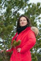 bonito morena mujer con largo Rizado pelo vestido en rojo invierno chaqueta y negro bufanda alrededor cuello, en pie en pino bosque foto