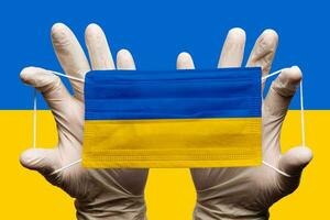 Doctor holding in white gloves protection medical face mask, respiratory bandage with National flag of Ukraine superimposed on mask. Concept photo