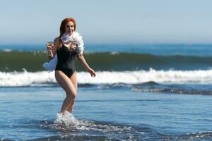 Smiling redhead woman in black swimsuit stands ankle deep in waves of ocean, holding glass of wine photo