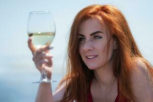 Closeup portrait happy redhead woman raised glass of wine at eye level and is looking intently at it photo