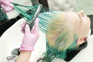 Hairdresser in protective glove holding wet hair in hand and brushes long green and bleached hair of customer while washing head in shower photo