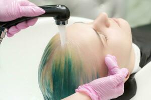 Hairstylist hands wash long hair of customer with green and discolored hair in sink with water from shower photo