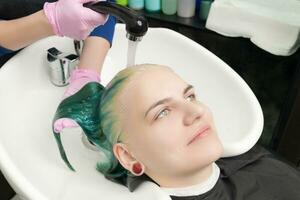 Hairstylist hands in glove washing green hair of young woman with shampoo in shower in special sink photo
