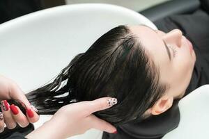 Hairdresser's hands wash long hair of brunette woman with shampoo in special sink for shampooing photo