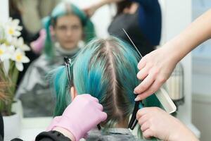Back view of female head with emerald hair color and regrown hair roots. Woman sits in chair by mirror, two hairdressers combing client's hair photo