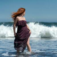 Woman in dress standing ankle deep in water on beach and turned head, looking over shoulder to sea photo