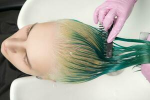 parte superior ver de estilista sostiene mojado pelo en mano y peines largo verde y descolorado pelo de cliente mientras Lavado pelo en ducha en especial lavabo foto