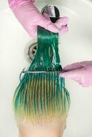 Angulo alto Disparo de peluquero en guante sostiene mojado pelo en mano y peinada largo verde y descolorado pelo de mujer mientras Lavado pelo en lavabo foto