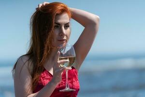 Portrait of redhead woman in dress holding glass of white wine at beach party during summer holidays photo