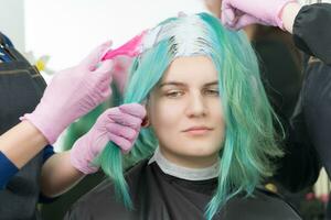 Process of bleaching hair roots in hair salon. Two hairdressers in glove use pink brush while applying paint to young adult customer with emerald hair color photo