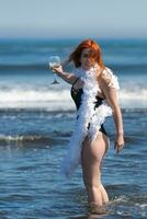 Smiling redhead woman in swimsuit stands ankle deep in breaking wave of sea, holding glass of wine photo