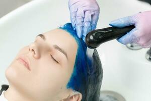 Hairdresser washes client's head with sapphire hair color after hair coloring process. Washing hair with shampoo in sink with special shower in beauty salon photo