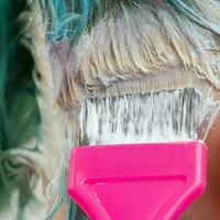 Hairdresser using pink brush while applying paint to female customer with emerald hair color during process of bleaching hair roots photo