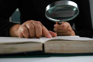 de cerca de un mujer mirando mediante un aumentador vaso a un libro de texto. aumentador vaso en mano y abierto libro en mesa. educación y investigación concepto. foto