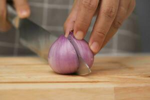 Female hand using knife to slice fresh red onion on chopping board, close-up. Chef chopping shallots using a sharp knife on a wooden chopping board. Preparing homemade food photo