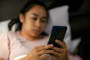 Young Asian woman lying on white bed and playing smartphone at night. Woman in bed with phone. Health and social concept. photo
