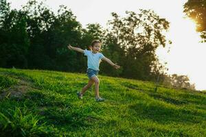 contento pequeño niña corriendo en el césped en el verano parque. pequeño niña corriendo mediante el prado con puesta de sol ligero. foto