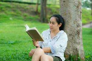 Cheerful young woman in jacket reading a book in summer park. Concentrated woman sitting on grass and studying under tree during vacation. Education concept photo