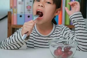 pequeño niña comiendo Fresco rojo uvas a hogar en el vivo habitación. linda joven asiático niña come sano frutas y Leche para su comida. sano comida en infancia foto