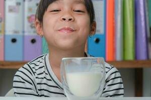 linda asiático niña Bebiendo un vaso de Leche a hogar en vivo habitación. pequeño niña Bebiendo Leche en el Mañana antes de yendo a escuela. sano comida en infancia. foto