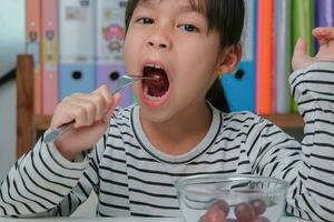 pequeño niña comiendo Fresco rojo uvas a hogar en el vivo habitación. linda joven asiático niña come sano frutas y Leche para su comida. sano comida en infancia foto