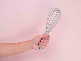 Close up of hand holding plastic whisk isolated on pink background. Kitchen tool for whipping cream or eggs. photo