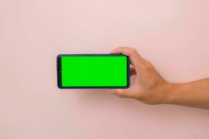 Human hand holding mobile smartphone with green screen in horizontal position isolated on pink background. photo