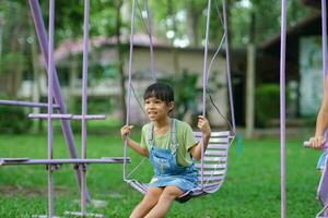 contento asiático niña teniendo divertido jugando en el patio de recreo durante verano. linda pequeño niña balanceo en el patio de recreo con un sonrisa y risa. activo verano ocio para niños. foto