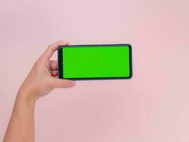 Human hand holding mobile smartphone with green screen in horizontal position isolated on pink background. clipping path photo