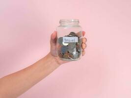 Close-up of young woman's hand holding glass jar with money  inside on pink background. Hand holding a money jar, travel, savings, education, donation. Finance plan concept. photo