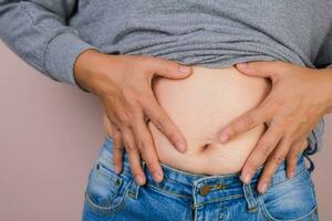 Fat woman hand holding excessive belly fat isolated on pink background. Overweight fatty belly of woman. Female diet and body health care concept photo