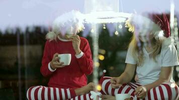 two kids in pajamas sitting on a wooden floor near the window with christmas light video