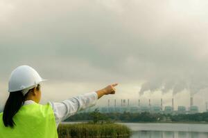 Female chief engineer wearing a green vest and helmet stands outside against the background of coal power plant station and steam in the morning mist. Engineer working and laptop at coal power plant. photo