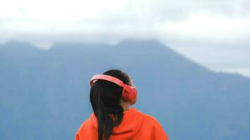 vista trasera de una mujer joven parada en una colina tranquila y escuchando música con auriculares por la mañana. mujer con un suéter disfrutando de la belleza de la naturaleza mirando la montaña en invierno. foto