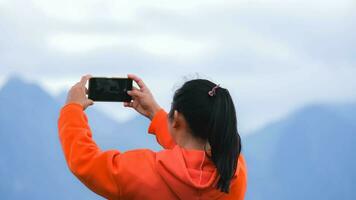 posterior ver de un joven mujer en pie en el parte superior de un calma montaña y tomando imágenes de el hermosa montaña paisaje a amanecer en el teléfono. foto