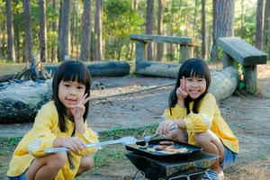 Happy family, cute sisters sitting on picnic by stove near tent and barbecuing in pine forest. Happy family on vacation in nature. photo