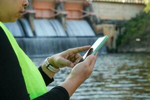 Female engineer in white hat working with smartphone and looking away at dam with hydroelectric power plant and irrigation. photo