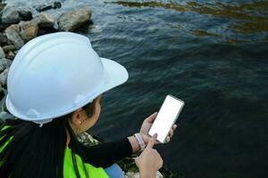 Environmental engineer uses a mobile phone to record water analysis data in dam. Environmentalists collect water samples from the dam to check for contamination. Water and ecology concept photo