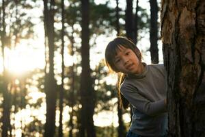 Funny little girl peeking out from behind tree in winter park. Cute smiling child playing hide and seek in the forest. Children look into the camera and smile. Happy family on vacation in nature. photo