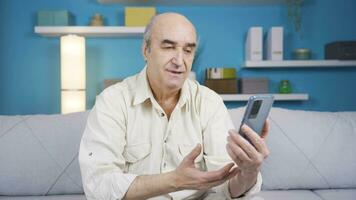 Smiling old man talking facetime on smartphone on sofa. video