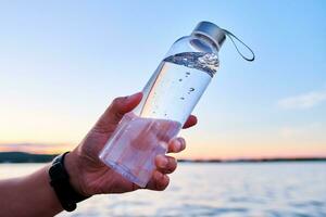 A man holds a bottle of water against the backdrop of a lake. photo