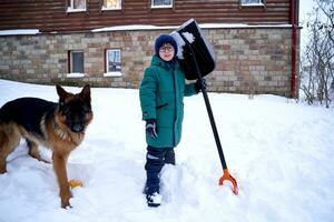 A boy in a green down jacket with a shovel stands in the yard in winter. photo