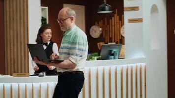 Senior pessoas dentro hotel recepção salão, mulher A chegar às frente escrivaninha com bagagem enquanto homem é esperando dentro salão área usando computador portátil. recepcionista ajudando Viajantes às luxo recorrer. portátil tomada. video