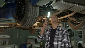 Handsome man examining lifted car with a lamp at the workshop video