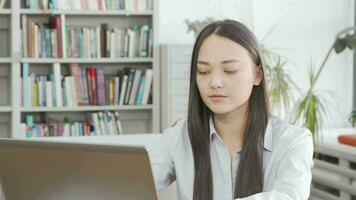 asiatisk ung kvinna leende till de kamera medan använder sig av bärbar dator på bibliotek video