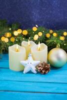 Christmas candles with fire, white sparkling star, golden pine cone and ball on blue wooden background. Behind there are spruce branches with defocused light. New Year. Copy space photo