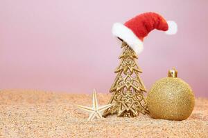 Golden Christmas tree in Santa's hat with shiny ball and starfish on sand of beach. New Year in hot countries. Copy space photo